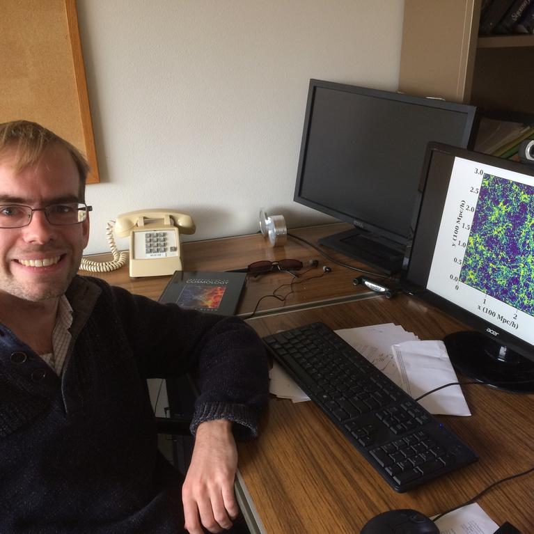 Dr. Simeon Bird poses, smiling in front of his computer